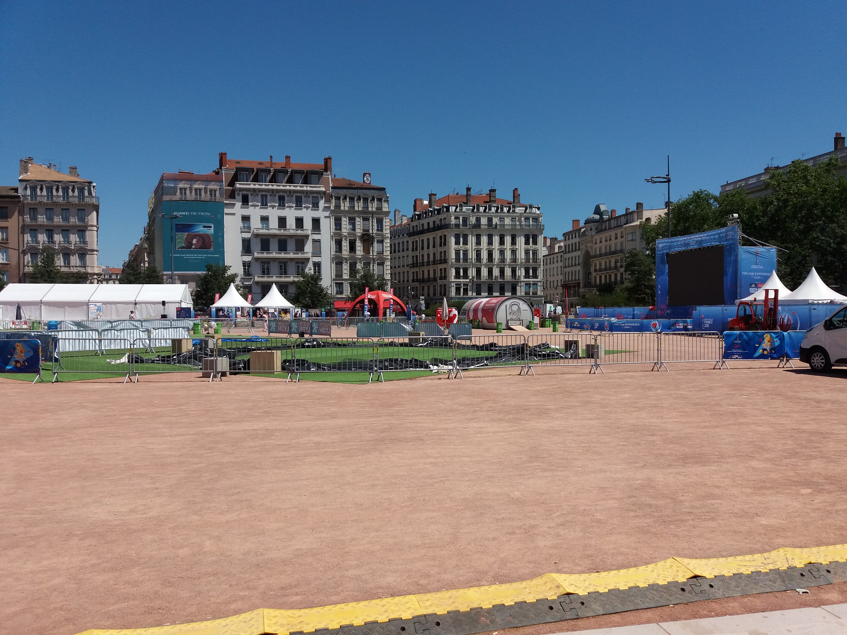 Großer Platz mit zahlreichen Zelten in der Mitte, umgeben von Gebäuden, Bäumen, Fahrzeugen auf der Straße und einem klaren blauen Himmel, umschlossen von einem Geländer.