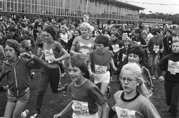 Schwarzes Foto von Kindern, die auf einem Rasenfeld bei einem Lauf mit einem Gebäude, Bäumen, Pfählen und einem klaren Himmel im Hintergrund laufen.