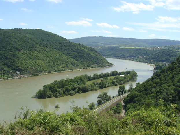 Eine malerische Aussicht auf den Rhein, der sich durch eine grüne Landschaft schlängelt, von einem Hügel aus, mit einer Straße daneben und weißen Wolken am Himmel.