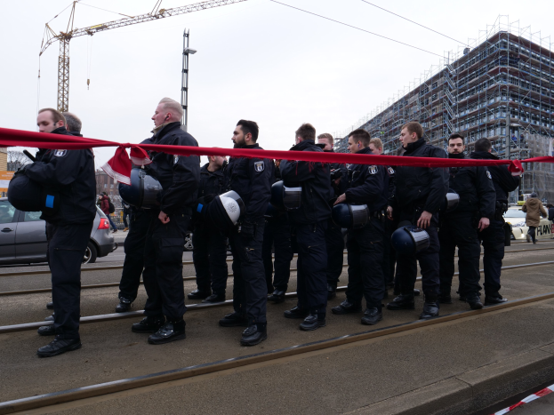 Eine Gruppe von Polizisten in schwarzen Uniformen und Helmen steht in einer Reihe auf einem Bahngleis mit einem roten Band davor, mit Fahrzeugen, Passanten, Gebäuden und einem klaren Himmel im Hintergrund.