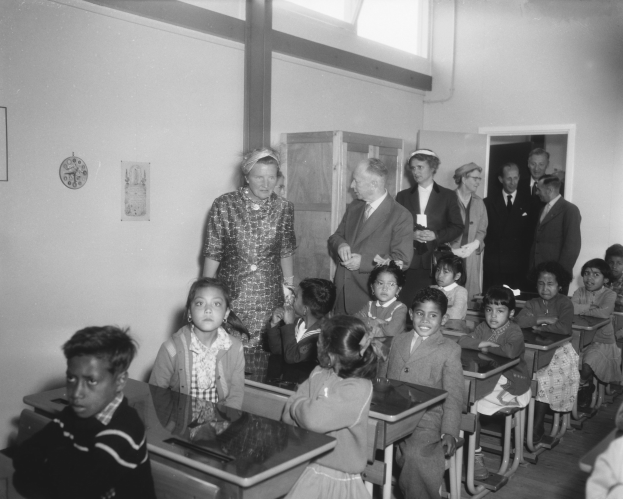 Schwarzes und weißes Foto einer Klasse mit Kindern auf Bänken sitzend und stehend, eine Frau in der Mitte, eine Uhr an der Rückwand, Papiere auf einem Schrank, Tür und Decke, sowie Deckenlampen.