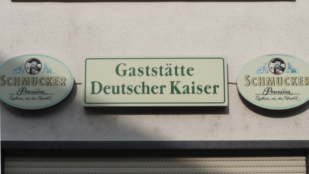 Drei hölzerne Bretter mit der Aufschrift "Gaststätte Deutscher Kaiser Schmucker" an einer Gebäudewand angebracht, mit einem Rollladen am Boden sichtbar.