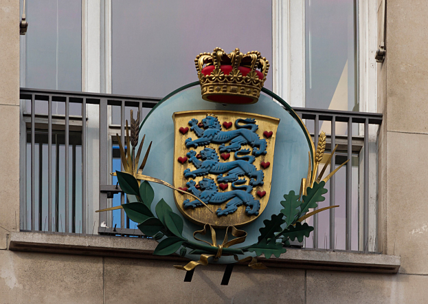 Wappen der britischen Königsfamilie an einer Gebäudewand mit einer Krone und einem Blatt-Logo, Fenstern und Metallgittern im Hintergrund.