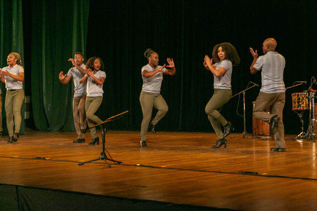 Eine Gruppe von Menschen auf einer Bühne mit Mikrofonen, Musikinstrumenten und einem Vorhang im Hintergrund, die Hände klappend, feiernd, wie eine Tanzmannschaft aussehend.