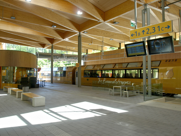 Ein belebter Bahnhof mit einem Zug auf den Schienen, umgeben von Bänken, Tischen, Mästen, Schildern, Lämpchen und Bäumen, mit Menschen, die auf den Zug warten.