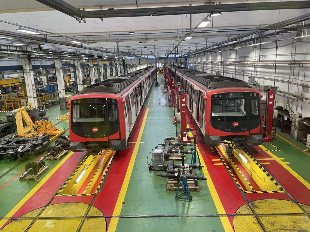 Zwei Züge auf den Schienen in einer Daimler Group-Fabrik, umgeben von Maschinen und Equipment, mit Rohren und Lichtern an der rechten Wand.