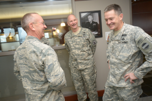 Drei uniformierte Männer, die vor einer Wand mit einem Foto Rahmen, einem Glasfenster mit Laternen und Tischen und einer braunen Tür auf der rechten Seite stehen und lächeln.