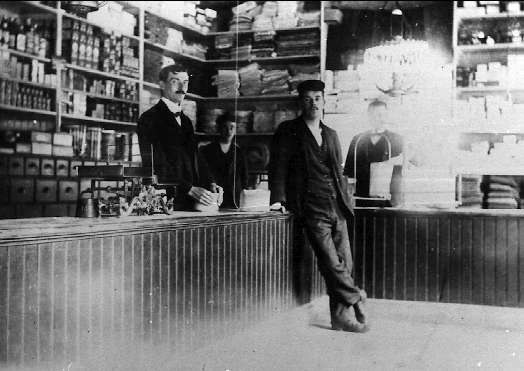 Schwarzes und weißes Foto von drei Männern an einer Ladentheke mit Gegenständen, einem Buchregal im Hintergrund und einer "The New York Times"-überschrift, die auf einen Raub hindeutet.