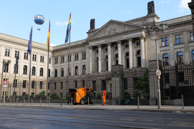Großes Gebäude mit vielen Fenstern, identifiziert als der Bundestag in Berlin, Deutschland, umgeben von Straßeninfrastruktur, Fußgängern, Fahrzeugen, Flaggen und Bäumen unter einem bewölkten Himmel.