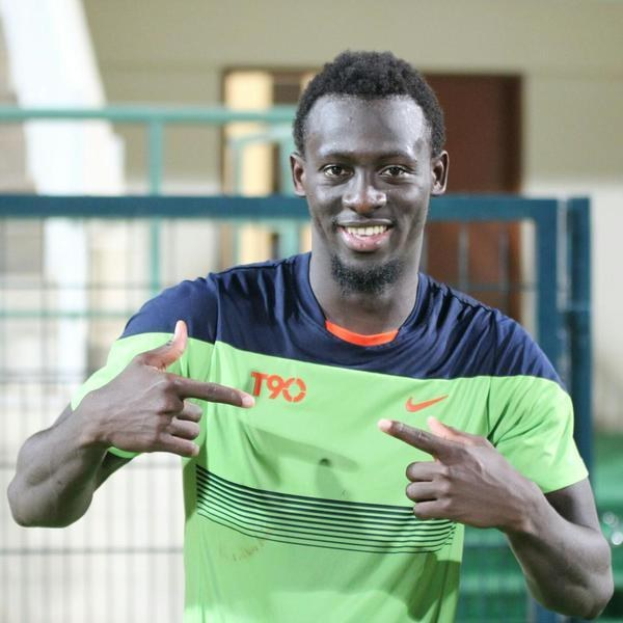 Ein Mann in einem grünen und blauen Hemd zeigt grinsend auf die Kamera, mit einem unscharfen Geländer, einer Wand und einer Tür im Hintergrund.