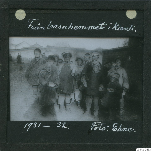 Schwarz-weiß-Foto einer Gruppe von Kindern, die zusammenstehen, einige halten Teller, mit Häusern im Hintergrund.