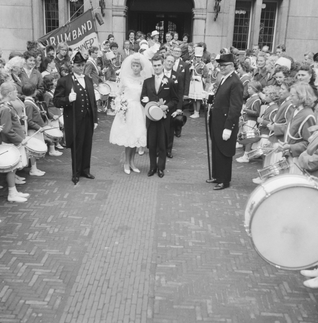 Schwarzes und weißes Foto einer Braut in einem weißen Kleid und Schleier und eines Bräutigams mit einem Blumenstrauß, der eine Straße entlang geht, vorbei an einer Menge mit einigen, die Musikinstrumente halten, vor einem Gebäude mit einer Fahne.