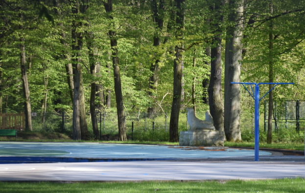 Basketballplatz in einem Park umgeben von Bäumen, mit einer Bank links, einem Zaun im Hintergrund und Gras unten.