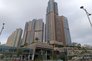 Eine Stadtstraße mit hohen Gebäuden, Fahrzeugen, Laternenmasten, einer Brücke, Bäumen und einem bewölkten Himmel, mit einem prominenten Wohnhaus im Vordergrund.