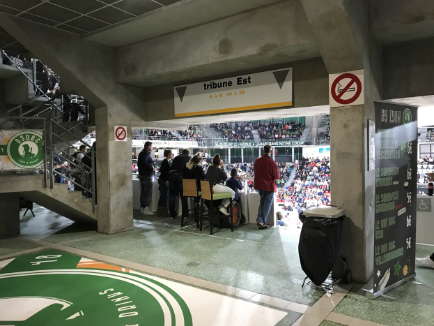Eine Gruppe von Menschen steht auf einem Stadionboden vor sitzenden Zuschauern, mit Schildern, die "Boston Celtics vs New York Jets" anzeigen, einem Müllcontainer, einer Tafel mit Text, einer Treppe mit Geländern und Deckenlampen unter einer Dachkonstruktion.