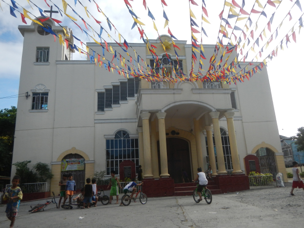 Menschen auf Fahrrädern vor einer weißen Kirche mit architektonischen Details, umgeben von Grün und einem bewölkten Himmel, mit bunten Fahnen darüber.