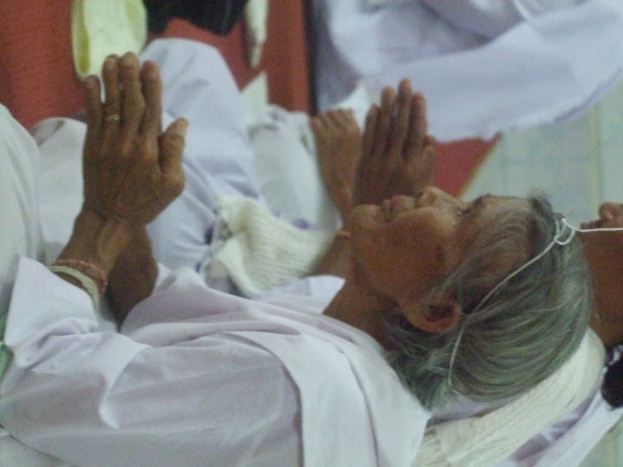 Eine ältere Frau liegt in einem Krankenhausbett, die Hände gefaltet, die Augen geschlossen und den Kopf auf den Händen ruhend, umgeben von ein paar Menschen im Hintergrund, die besorgt aussehen.