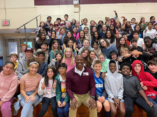 Schüler posieren für ein Foto in einer Turnhalle mit einigen auf der Treppe sitzend und anderen stehend, eine Wand im Hintergrund, eine Tür und ein Ausgangsschild links.