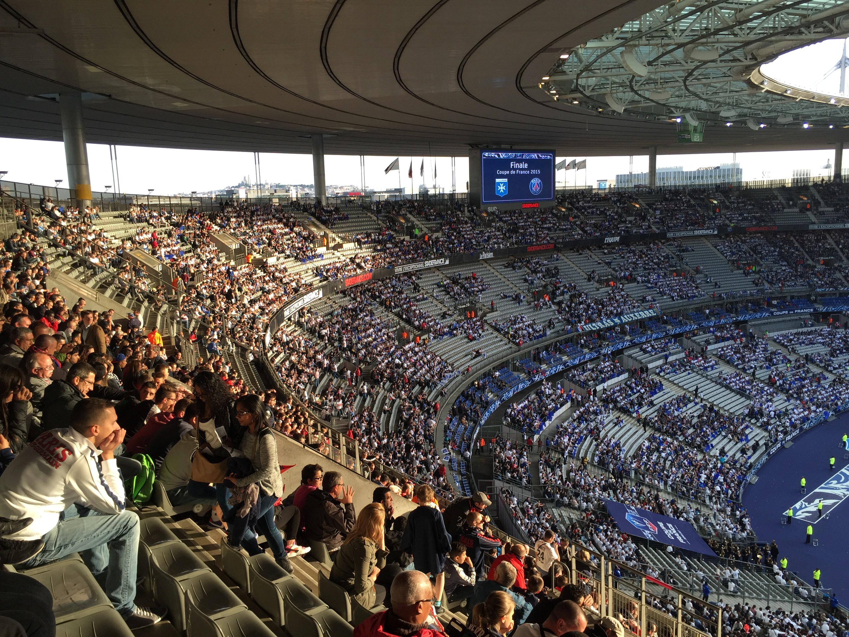 Großes Publikum in einem Stadion bei einem Fußballspiel mit einer Bühne, Fahnen, Stangen, einem Bildschirm und der Allianz Arena in München, Deutschland im Hintergrund.