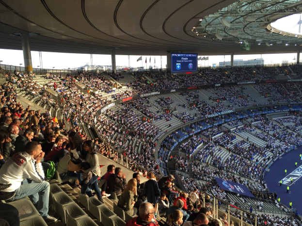 Großes Publikum in einem Stadion bei einem Fußballspiel mit einer Bühne, Fahnen, Stangen, einem Bildschirm und der Allianz Arena in München, Deutschland im Hintergrund.