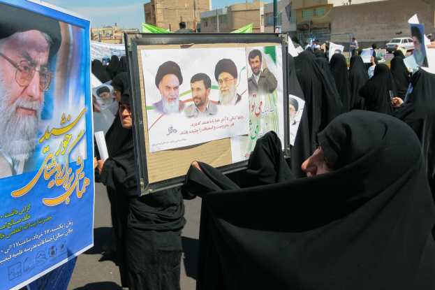 Eine Gruppe von Frauen in schwarzen Abayas hält Plakate mit Bildern von Ajatollah Ali Khamenei hoch, im Hintergrund sind Gebäude, Pfosten, Fahrzeuge und ein klarer blauer Himmel zu sehen.