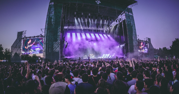 Menschenmenge auf einem Musikfestival mit Künstlern auf der Bühne unter Scheinwerfern, umgeben von Bildschirmen, mit einem Gebäude, Bäumen und blauem Himmel im Hintergrund.