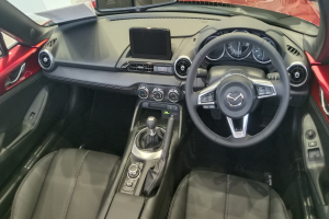 Das Innere eines roten Mazda MX-5 Miata RF mit schwarzen Ledersitzen, einem Lenkrad, einem Schaltknüppel, Knöpfen, einem Bildschirm und einem Rückspiegel.