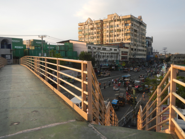 Eine Fußgängerbrücke mit Geländern überspannt eine vielbefahrene Straße mit vielen Fahrzeugen und Fußgängern, mit Gebäuden, Bäumen, Strommasten mit Drähten und einem bewölkten Himmel im Hintergrund.
