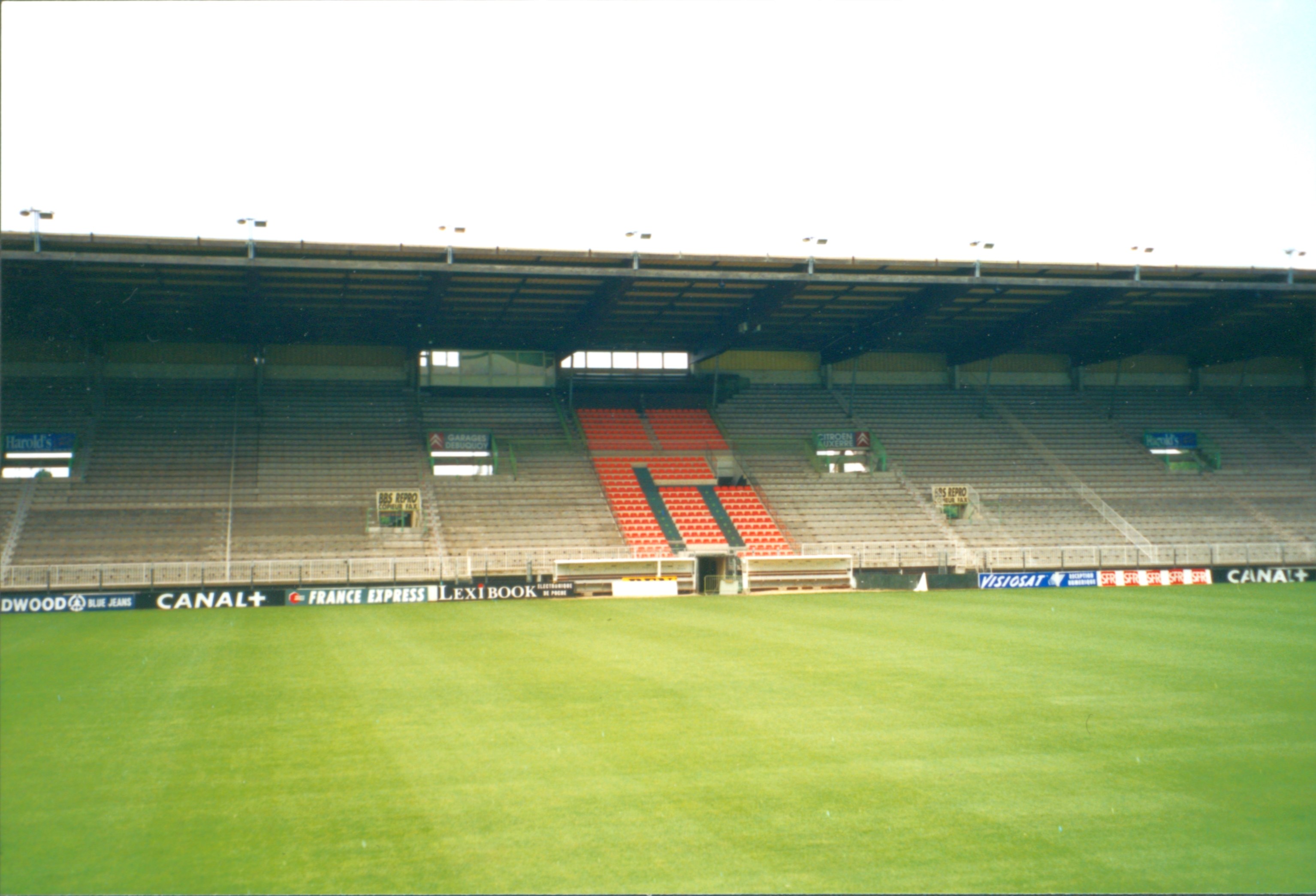 Fußballfeld mit grünem Rasen, einem großen leeren Stadion im Hintergrund und einem klaren blauen Himmel darüber.