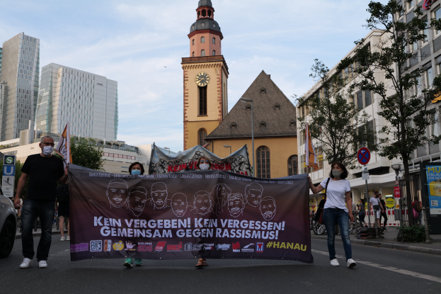 Eine Gruppe von maskierten Menschen, die eine Straße entlanggehen und ein Banner halten, mit einem geparkten Auto auf der linken Seite, Gebäuden, Bäumen, Schildern, Pfählen, einem Uhrenturm und einem klaren blauen Himmel im Hintergrund.
