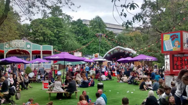 Große Gruppe von Menschen auf einem Rasenfeld mit Schirmen, Tischen und einer Tafel, die ein Biergarten in London anzeigt.