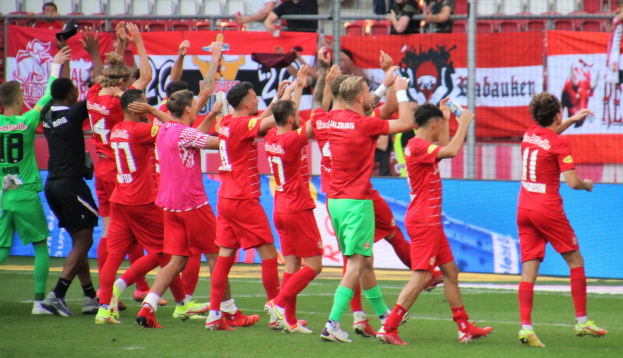 Gruppe von Fußballspielern, die ein Tor feiern, mit Flaschen oder klatschenden Händen, sowie Bannern, einem Zaun, Stühlen und einem Dach mit Lampen im Hintergrund.