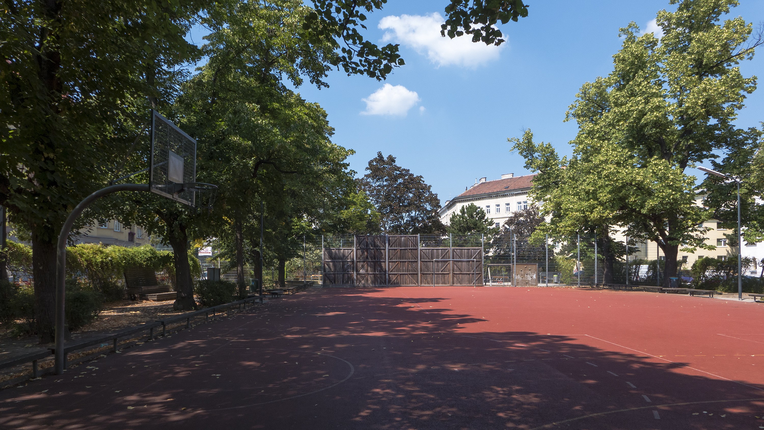 Ein Basketballplatz in einem Park umgeben von Bäumen, Gebäuden, Straßenlaternen, Grills, Bänken, Pflanzen und einem bewölkten Himmel.