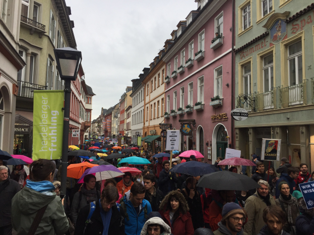 Große Gruppe von Menschen, die mit Schirmen auf einer Straße gehen, einige tragen Taschen und halten Schilder, mit Gebäuden und einem Lichtmast im Hintergrund.