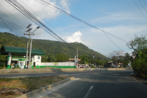 Tankstelle mit elektrischen Masten, Kabeln, Namensschildern, Bäumen, Gebäuden, Fahrzeugen, Hügeln und bewölktem Himmel im Hintergrund.