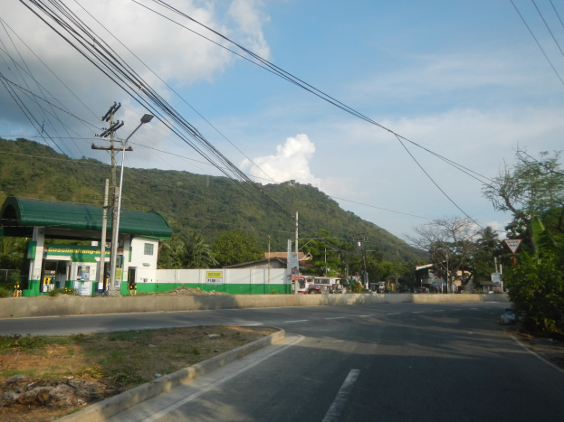 Tankstelle mit elektrischen Masten, Kabeln, Namensschildern, Bäumen, Gebäuden, Fahrzeugen, Hügeln und bewölktem Himmel im Hintergrund.