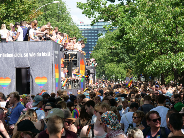Große Menschenmenge vor Lastwagen bei der Christopher Street Day Parade in Berlin, viele mit Mützen und Schutzbrillen, einige mit Fahnen, mit einem Banner auf dem Lastwagen und Bäumen, Gebäuden und einem Laternenpfahl im Hintergrund bei bewölktem Himmel.