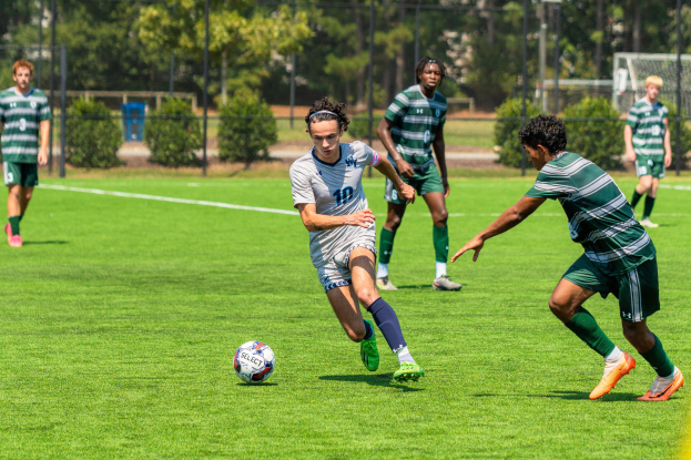 Junge Männer in Sportuniformen spielen Fussball auf einem Rasenfeld mit Bäumen und einem Tor im Hintergrund.