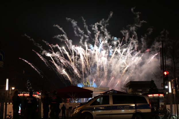 Polizeiauto vor einer Menschenmenge bei einer Silvesterfeier in Berlin geparkt, mit farbigen Feuerwerken am Himmel und Stadtlichtern.