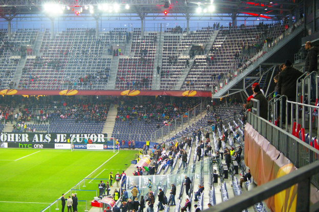 Großes Stadion voller Zuschauer bei einem Fußballspiel, mit sitzenden und stehenden Personen, einem Geländer, einer Fahne und einem leeren Abschnitt mit Deckenbeleuchtung.