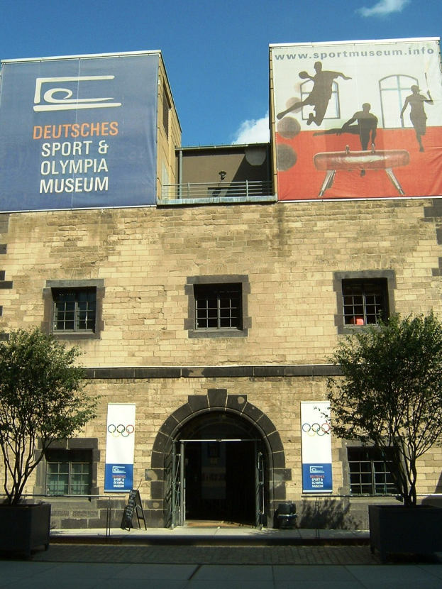 Außenansicht des Deutschen Sport & Olympia Museums in Berlin mit einem Gebäude mit Fenstern und einer Tür, umgeben von Bäumen und Bannern unter einem bewölkten Himmel.