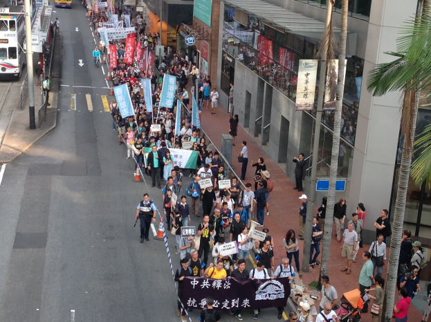 Eine große Gruppe von Menschen marschiert auf einer Straße in Hong Kong, mit Protestschildern in den Händen, Bäumen und glasverkleideten Gebäuden auf der rechten Seite und Fahrzeugen auf der linken Seite.
