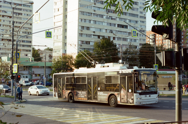 Stadtstraße mit einem Bus, der an hohen Gebäuden, Bäumen, Laternen, Ampeln, Schildern, Fußgängern auf dem Gehweg, einem Geländer und einem klaren blauen Himmel vorbeifährt.