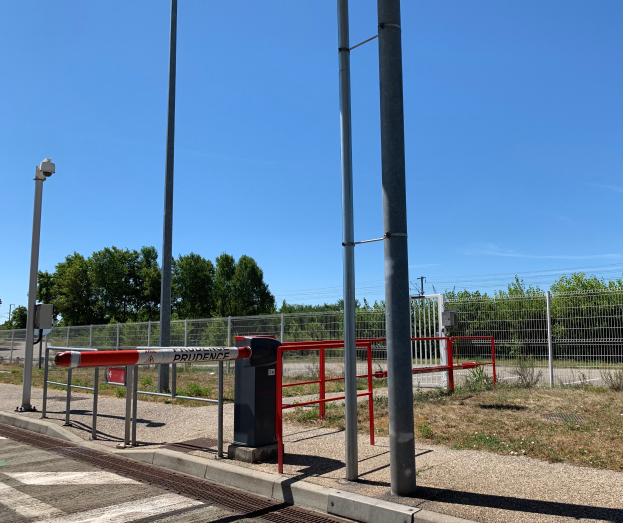 Ein Bahnhofsgebäude mit einem roten und weißen Schild, umgeben von Pfählen, einem Metallzaun, Bäumen, Pflanzen, Gras, einem Straßenpfahl, Drähten und einem bewölkten Himmel, mit einer zentralen Absperrung, um den Zugang zu verhindern.