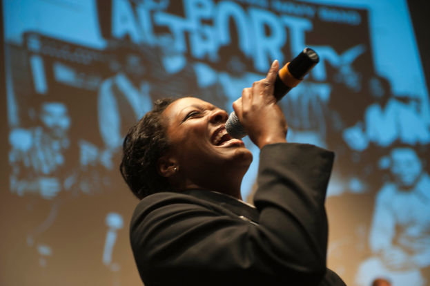 Eine Frau in einem schwarzen Kleid singt in ein Mikrofon auf der Bühne, ihre Augen geschlossen und ihr Gesicht von Scheinwerferlicht erhellt.