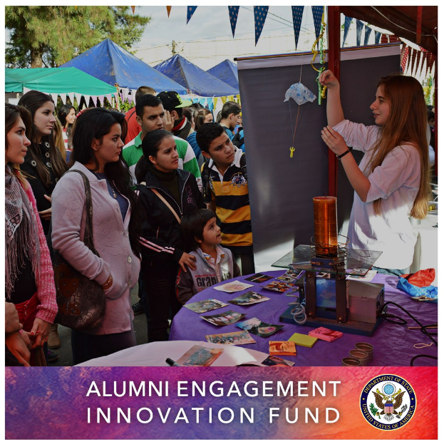 Gruppe von Menschen um einen Tisch versammelt mit einer "Alumni Engagement Innovation Fund"-Aufschrift, umgeben von Gegenständen, mit Zelten, Bäumen und einem klaren blauen Himmel im Hintergrund und Text und Logo unten.