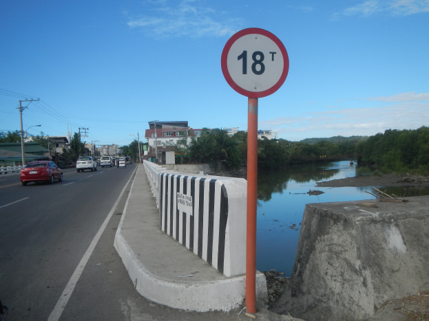 Ein Tempolimit-Schild am Straßenrand neben einem Fluss, mit Fahrzeugen, einer Barriere, Bäumen, Gebäuden, Strommasten mit Drähten und einem bewölkten Himmel im Hintergrund.