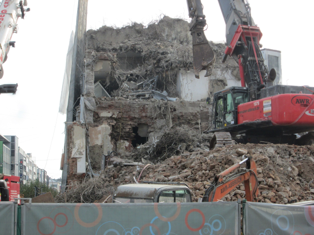 Ein Kran im Vordergrund zerstört ein Gebäude, umgeben von anderen Kränen, Fahrzeugen, Felsen und einem Banner mit Text, mit Gebäuden, Bäumen und dem Himmel im Hintergrund.