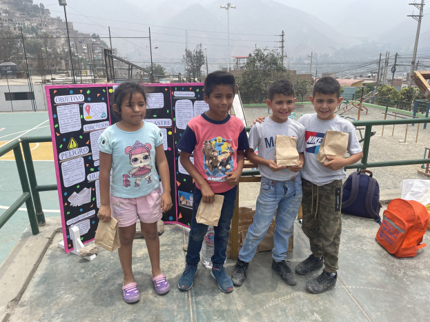Eine Gruppe von Kindern steht auf einem Basketballfeld und hält Papiertüten in den Händen, mit Tüten und Gegenständen rechts daneben und einer Tafel mit Text links daneben, vor dem Hintergrund von Zäunen, Pfosten, Straßenlaternen, Bäumen, Gebäuden, Hügeln und dem Himmel.
