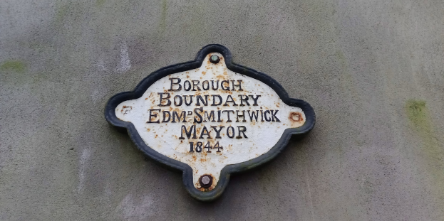 Metallplakette an einer Gebäudeseite mit schwarzem Hintergrund und weißer fettgedruckter Schrift, die "Stadtteilgrenze Edm Smithwick Bürgermeister" liest.